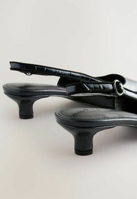 Musta slingback-korkokengät kiiltävällä pinnalla ja kohtuullisilla koroilla, aseteltuna vaalealla taustalla, näyttäen suunnittelun yksityiskohtia.