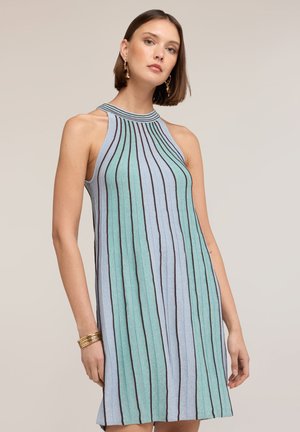 Kobieta w sukience bez rękawów z wysokim kołnierzem, z pionowymi niebieskimi i zielonymi paskami, złotymi kolczykami i wieloma złotymi bransoletkami, stojąca na jednolitym tle.