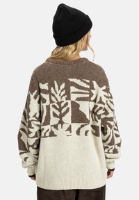 Vrouw in een beige en bruine gebreide trui met patroon en een zwarte beanie, van achteren gezien tegen een effen witte achtergrond.