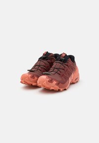 Pantofi de trail running cu un superior de culoare maro-roșiatică și talpă de un corai. Prezintă o suprafață texturată, sistem de șireturi integrat și cuie pentru aderență.