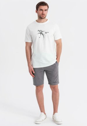 Hvid bomulds t-shirt med et cykelgrafik og tekst, parret med grå sportsshorts. Fodtøj er hvide sneakers. Enkel design.