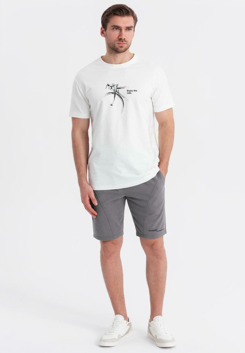 Vit bomull t-shirt med en cykelgrafik och text, kombinerad med grå sportshorts. Skorna är vita sneakers. Enkelt design.