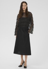 Sieviete, ģērbta melnā caurspīdīgā mežģīņu augšpusē ar garām piedurknēm, augsto jostasvietu melnā midi svārkos un melnās augstpapēžu kurpēs, stāv ar vienu roku kabatā.