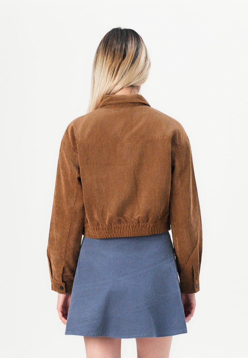 Personne avec de longs cheveux blonds portant une veste en velours côtelé marron, courte, et une mini-jupe bleue, se tenant de dos.