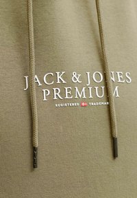 Khaki mikina s pletenými šňůrkami a stříbrnými kovovými konci. Na přední straně je bílý potisk s textem "JACK & JONES PREMIUM" a symbolem registrované ochranné známky.