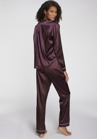 Satin pajama set in deep purple with a relaxed fit. Features a long-sleeve top and pants with white piping along the edges.