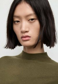 Primo piano di una persona con capelli neri lisci e trucco neutro, che indossa un top aderente verde oliva con un collo alto. Texture della pelle liscia.