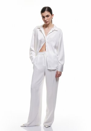 Witte satijnen blouse met een ontspannen pasvorm, voorzien van een puntige kraag, knoopsluiting aan de voorkant en open zijkanten. Gecombineerd met bijpassende wijde broek.