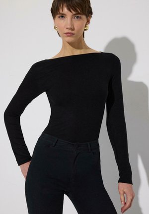 Νεαρή γυναίκα που φοράει μαύρο τοπ με μακριά μανίκια χωρίς ώμους και μαύρο παντελόνι, στέκεται με το ένα χέρι στη μέση, μπροστά σε απλό φόντο.