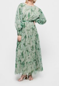 Light green maxi dress in sheer fabric with tiered layers, balloon sleeves, and a belted waist. Features abstract green patterns.