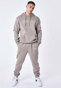 Conjunto de sudadera y pantalones de chándal en color beige claro con capucha. La sudadera tiene un bolsillo delantero y un logotipo blanco. Los pantalones de chándal cuentan con puños elásticos y cordón ajustable.