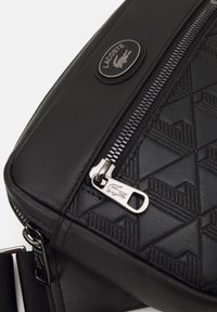 Czarna syntetyczna torba crossbody z geometrycznym teksturowanym wzorem, metalowym zamkiem błyskawicznym i okrąłym srebrnym akcentem logo Lacoste.