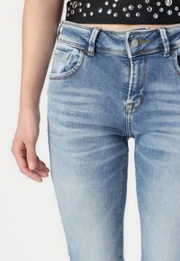 Jean en denim bleu clair avec une taille haute, dotés de poches avant, d'une texture décolorée et de coutures contrastées. Portés avec un haut noir.