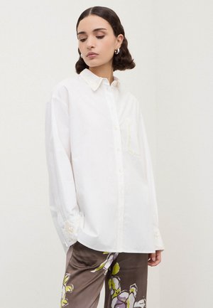 Donna con capelli corti e scuri che indossa una camicia bianca larga con bottoni e pantaloni marroni a fantasia floreale, guarda in basso di lato su uno sfondo uniforme.