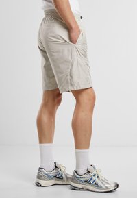 Beige Sport Shorts mit elastischem Bund, Seitentaschen und weißen Paspeln an den Nähten. Kombiniert mit silbernen Laufschuhen.