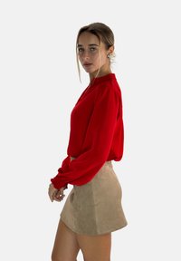 Red long-sleeve blouse with gathered cuffs, paired with a beige suede mini skirt. The blouse has a high neckline and a smooth texture.
