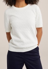 Pull blanc texturé à manches courtes, présentant un motif de points en relief et un col et un ourlet côtelés. Associé à un pantalon navy foncé.