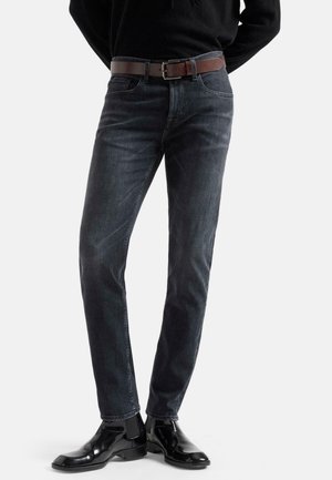 Man wearing slim-fit dark gray jeans with a brown belt and shiny black dress boots, standing with hands behind back.