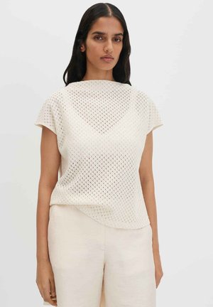 Femme aux longs cheveux foncés portant un haut en maille crème à manches courtes et un pantalon beige clair, debout devant un fond uni.