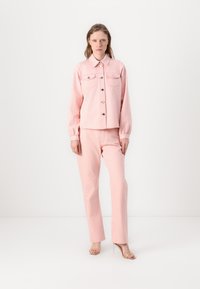 Pink denim jacket with front pockets and buttons, paired with matching pink trousers, both have a textured finish. Model wears clear heeled sandals.