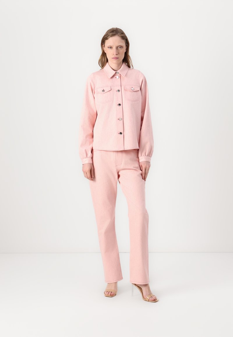 Pink denim jacket with front pockets and buttons, paired with matching pink trousers, both have a textured finish. Model wears clear heeled sandals.