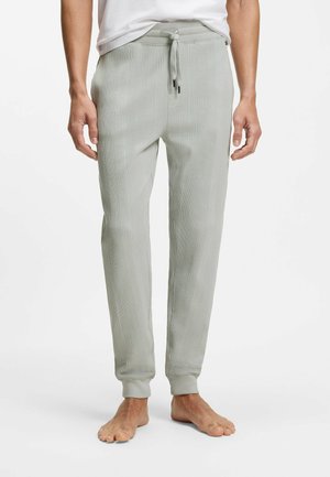 Mand iført lysegrå teksturerede strikkede joggingbukser med snøre i taljen og manchetter ved anklerne, stående barfodet mod hvid baggrund.