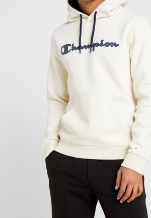 Hombre con sudadera crema de Champion con cordones azul marino y pantalones negros, con las manos en los bolsillos, cortada a la altura del cuello y los muslos.