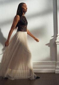 Schwarzer Spitzen-Body mit transparenter Textur, kombiniert mit einem fließenden beigen plissierten Rock. Das Model trägt schwarze High Heels und wird in sanftem natürlichen Licht präsentiert.