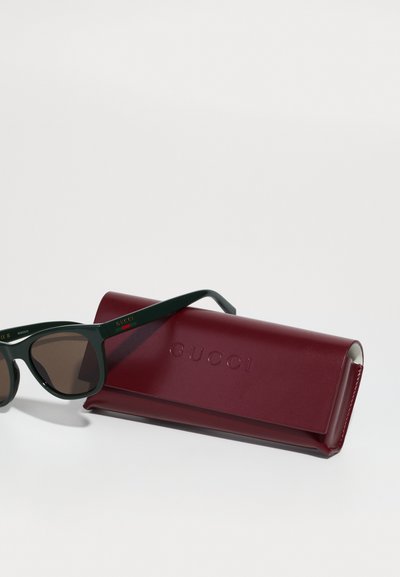 Melna un zaļa acetāta saulesbrilles ar brūnām lēcām, blakus burgundijas krāsas ādas maciņam ar izsistu uzrakstu "GUCCI".