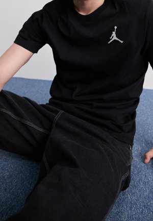 Czarny bawełniany t-shirt z małym szarym logo Jordana na klatce piersiowej, zestawiony z luźnymi czarnymi spodniami z białymi przeszyciami.