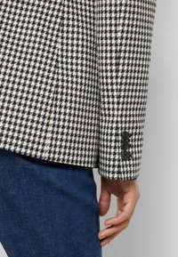 Blazer a quadri con un motivo houndstooth bianco e nero, tessuto morbido e tre bottoni neri sul polsino, abbinato a jeans blu scuro.