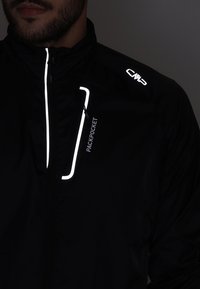 Veste coupe-vent noire avec des accents réfléchissants, dotée d'une poche frontale zippée étiquetée "PACKPOCKET" et d'un logo sur l'épaule.