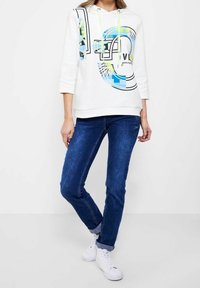 Sudadera blanca con un diseño gráfico, que presenta acentos en azul, negro y verde. Combinada con jeans azul oscuro y zapatillas blancas.