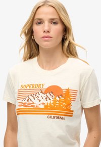 T-shirt crème avec un imprimé graphique représentant des montagnes, des arbres et un soleil dans des tons orangés. Manches courtes et encolure ronde.