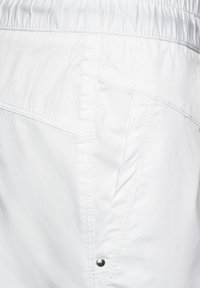 Närbild av vit tyg med diagonala sömmar, en ficka, elastisk midja och en liten silvernittingdetalj.