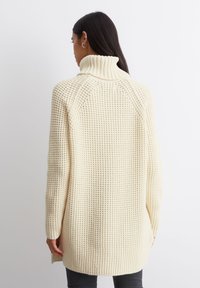 Beige strikkegenser med høy krage, med løs passform, teksturert overflate og ripede detaljer langs kragen og mansjettene.