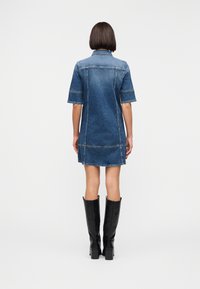 Γυναίκα που στέκετα ι με την πλάτη γυρισμένη, φορώντας ένα κοντομάνικο τζιν φόρεμα και μαύρες δερμάτινες μπότες μέχρι το γόνατο, σε απλό φόντο.