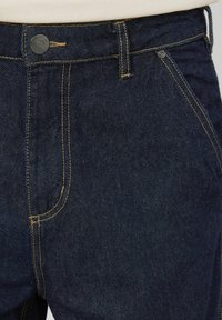 Jean en denim bleu foncé avec une coupe droite, des coutures jaunes contrastantes et une fermeture classique à bouton. Doté de poches avant angulaires.