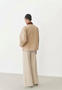 Person mit dunklem Haar im niedrigen Dutt, der beige weite Hosen und eine beige Jacke mit braunem Kragen trägt, steht vor einem schlichten hellen Hintergrund.