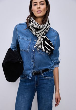 Frau trägt ein blaues Jeanshemd und Jeans, schwarzen Gürtel, Schal mit Tiermuster und hält eine schwarze Tasche vor einem einfarbigen Hintergrund.