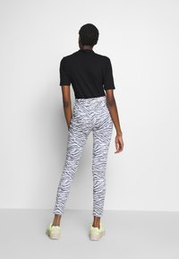 Persona con cabello corto y rizado lleva una camiseta negra de manga corta, pantalones de tiro alto con estampado de cebra en blanco y gris, y zapatillas beige con acentos neón.