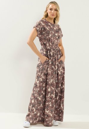 Frau trägt ein langes, ärmelloses Blumenmusterkleid mit Taschen und weißen Turnschuhen, steht vor einem schlichten hellen Hintergrund.