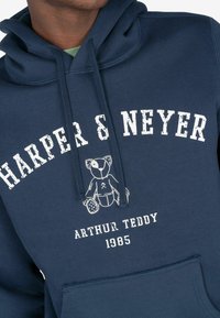 Granatowy hoodie z kieszenią kangurką, z białym napisem "HARPER & NEYER" oraz grafiką misia. Wykonany z miękkiego, teksturowanego materiału.