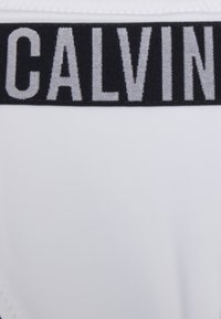 Biele plavky so čiernym pásom, na ktorom je veľkými, kontrastnými sivými písmenami napísané "CALVIN". Tkanina vyzerá hladko a elasticky.