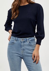 Pull en maille marine à manches longues bouffantes et col côtelé, associé à un jean en denim bleu clair taille haute avec cinq poches.