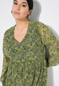 Abito floreale verde con scollatura a V, dotato di lunghe maniche trasparenti e vita arricciata. Il tessuto presenta motivi di fiori bianchi e gialli.