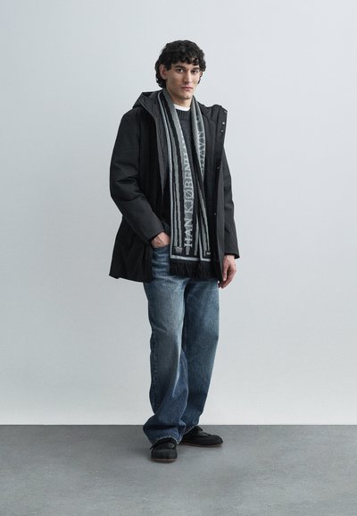 Jachetă impermeabilă neagră, pulover tricotat închis la culoare, eșarfă gri cu model, blugi evazați albaștri și încălțăminte neagră, stând pe o suprafață netedă gri.