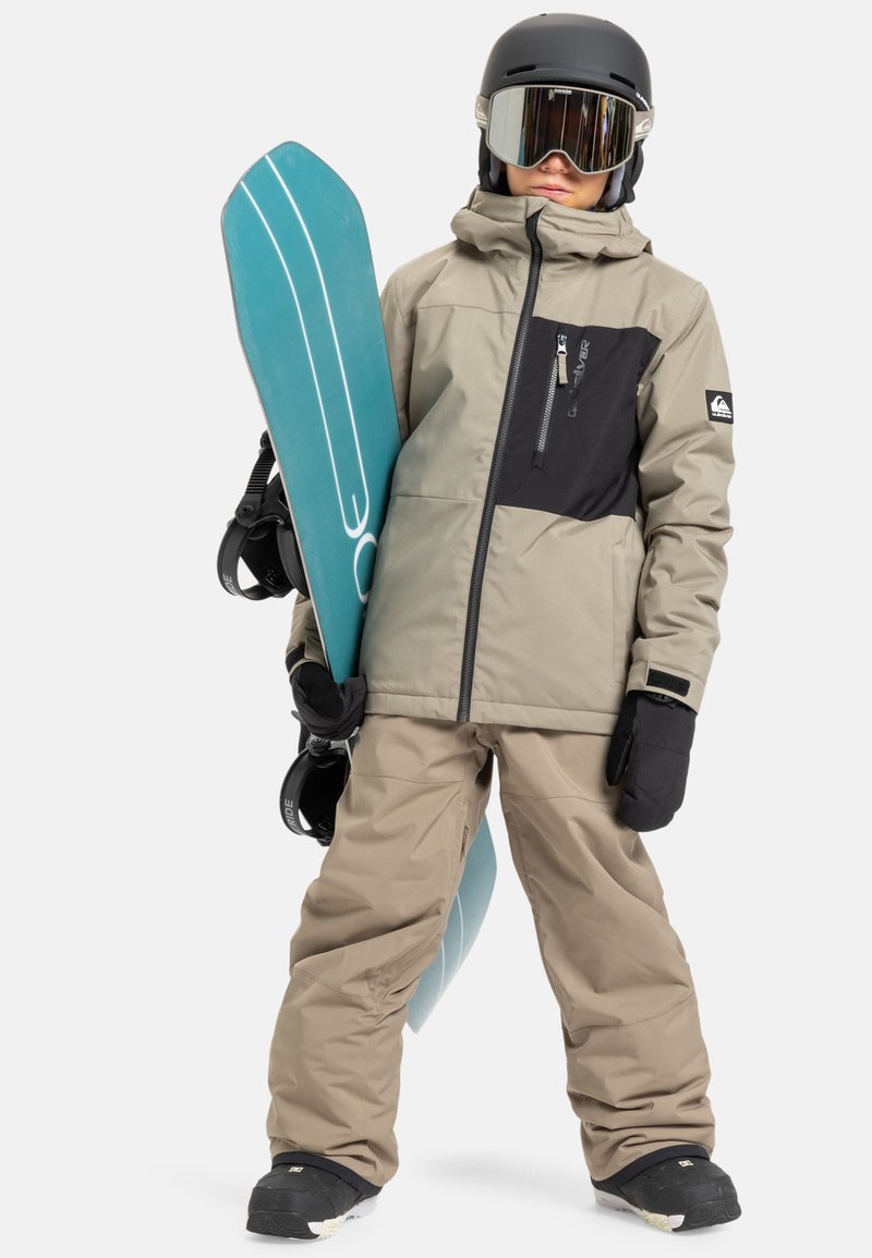 Osoba w beżowej kurtce i spodniach narciarskich, czarnym kasku, goglach i rękawicach, trzyma niebieską deskę snowboardową pionowo przy boku.