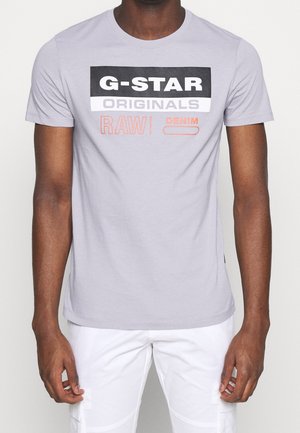 Homme portant un t-shirt à manches courtes gris clair avec le texte « G-STAR ORIGINALS RAW DENIM » et un pantalon cargo blanc, debout devant un fond blanc.