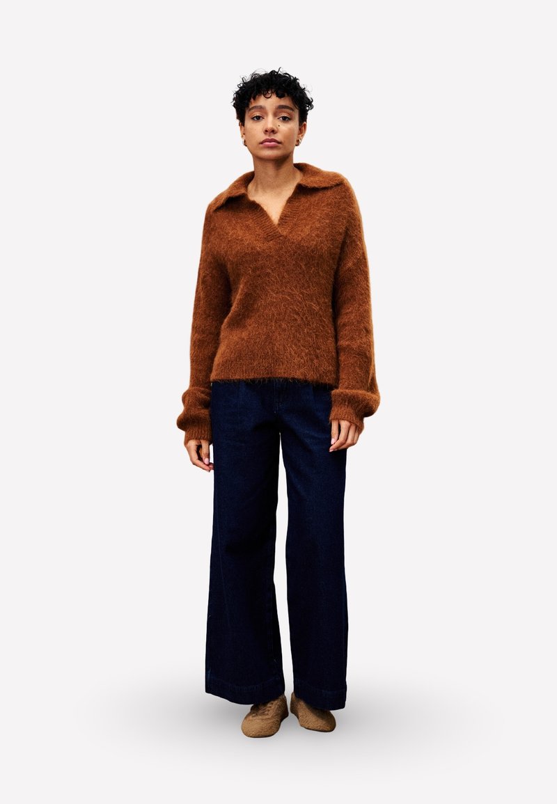 Brauner, flauschiger Pullover mit V-Ausschnitt und Kragen, kombiniert mit weit geschnittenen, dunkelblauen Jeans. Das Model steht vor einem neutralen Hintergrund.